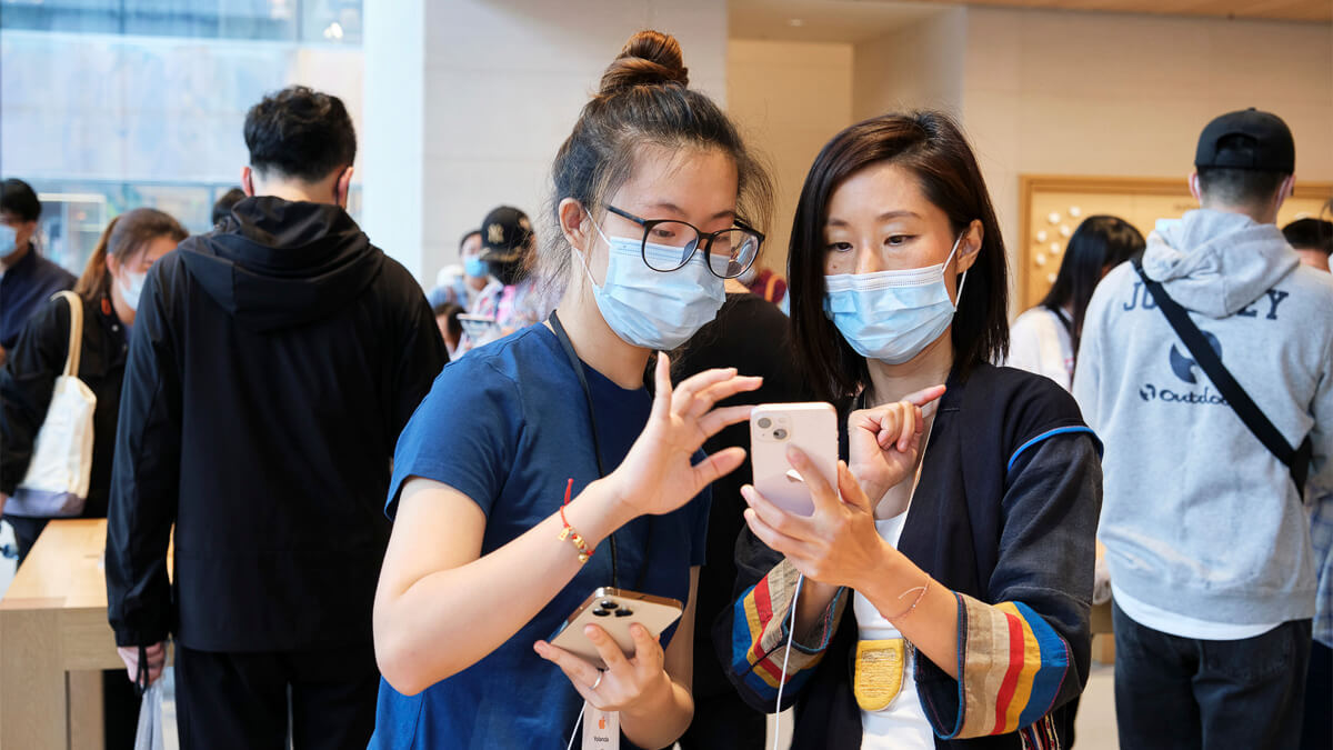 Two women wearing masks look at their smartphones together in a crowded indoor space.