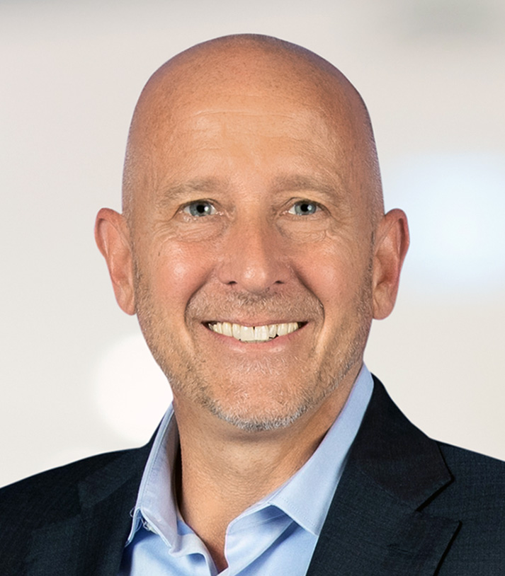 A smiling, bald man with blue eyes wearing a dark suit and light blue shirt against a neutral background.