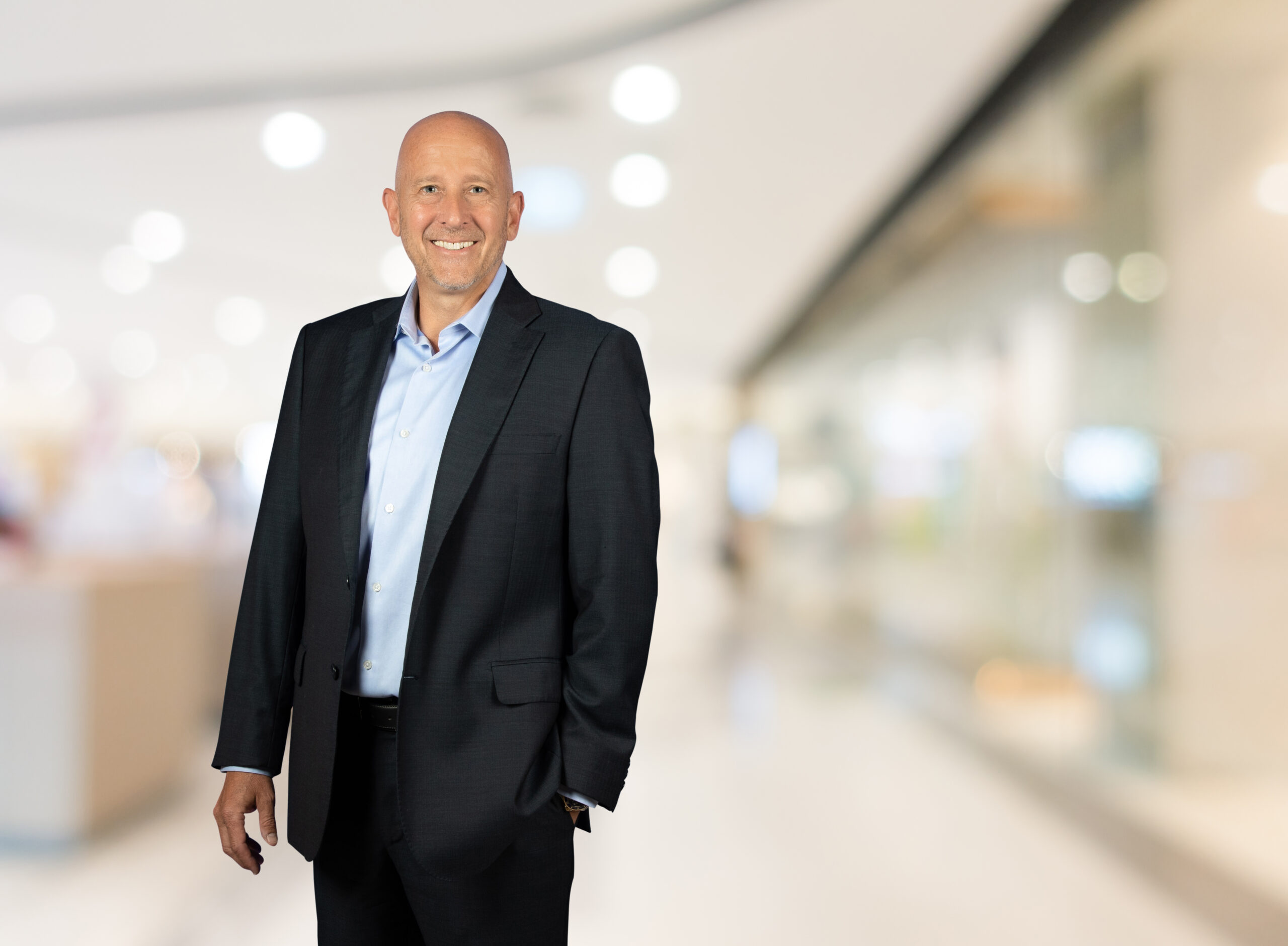 A smiling, bald man in a dark suit and light blue shirt stands confidently in a bright, blurred indoor setting.