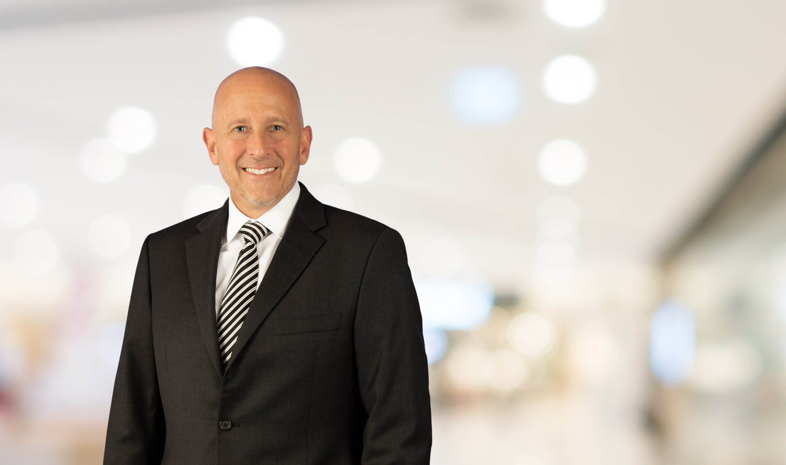 A smiling, bald man in a black suit, white shirt, and striped tie stands against a blurred indoor background.