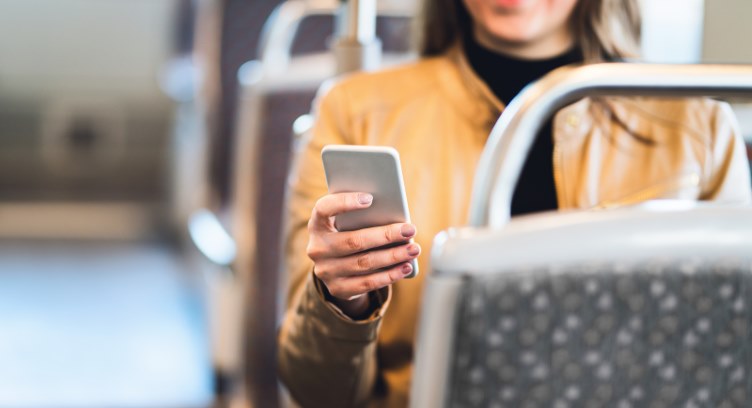Smiling woman in a tan jacket using her smartphone while seated on a train or bus.