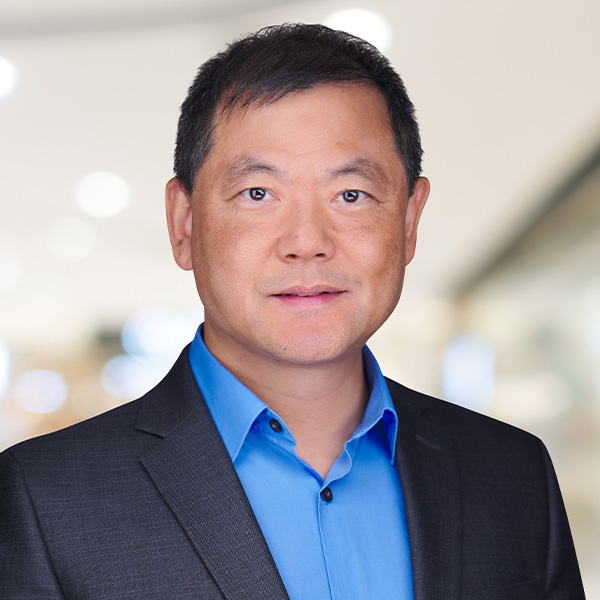 A professional Asian man in a dark suit and blue shirt, standing in a bright, blurred indoor setting.