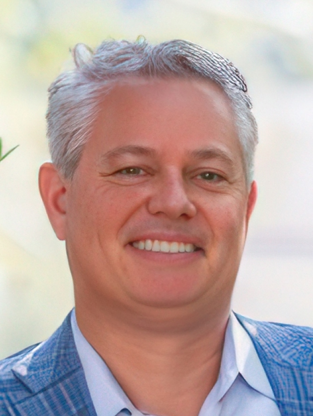 A smiling man with short gray hair wearing a blue checkered blazer and light blue shirt, standing outdoors.