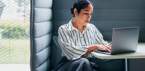 A woman in a striped shirt works on a laptop at a small table in a modern, cozy workspace.
