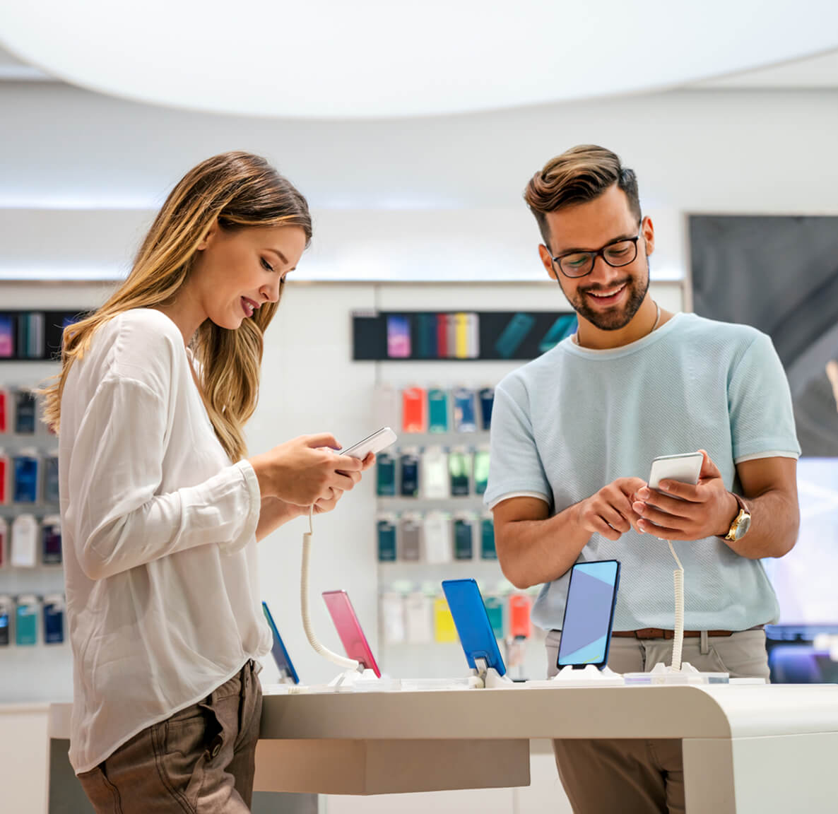 Two smiling customers try out smartphones at a store display with multiple devices on stands.