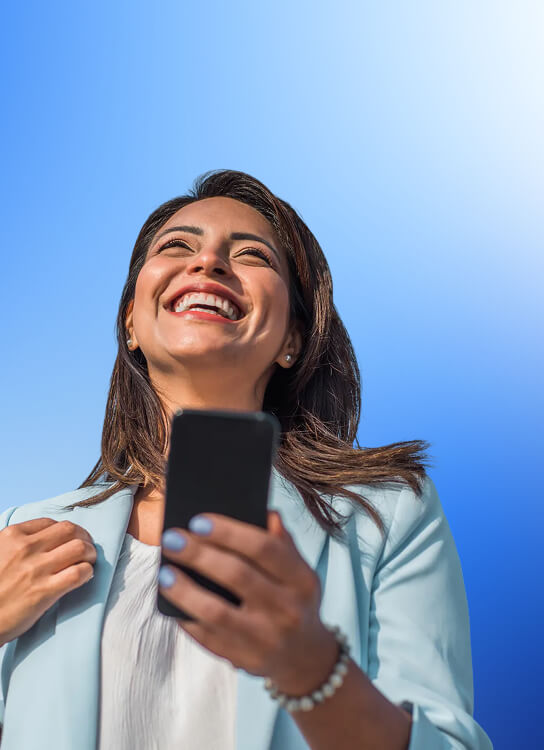 A woman smiling joyfully while looking at her smartphone against a bright blue sky background.