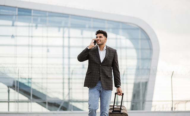A man with a suitcase talks on his phone outside a modern glass building, with a confident smile and casual attire.