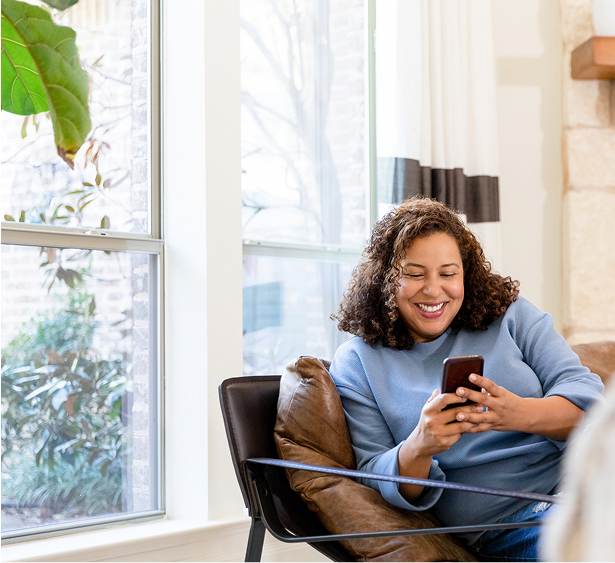 A woman with curly hair smiling while looking at her phone indoors near a large window.