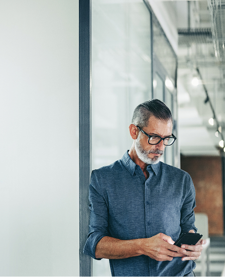 A mature man with glasses and a gray beard is looking at his phone while standing in a modern office.