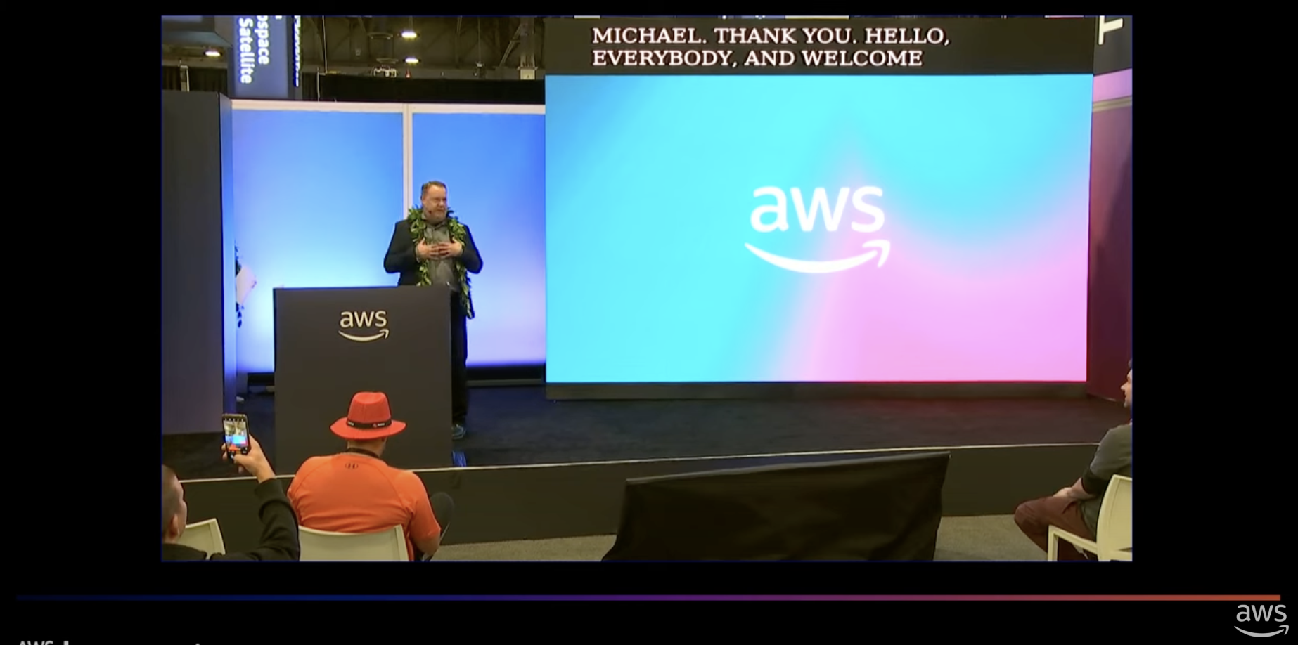 A man wearing a lei speaks on stage at an AWS event with a large screen displaying the AWS logo behind him.