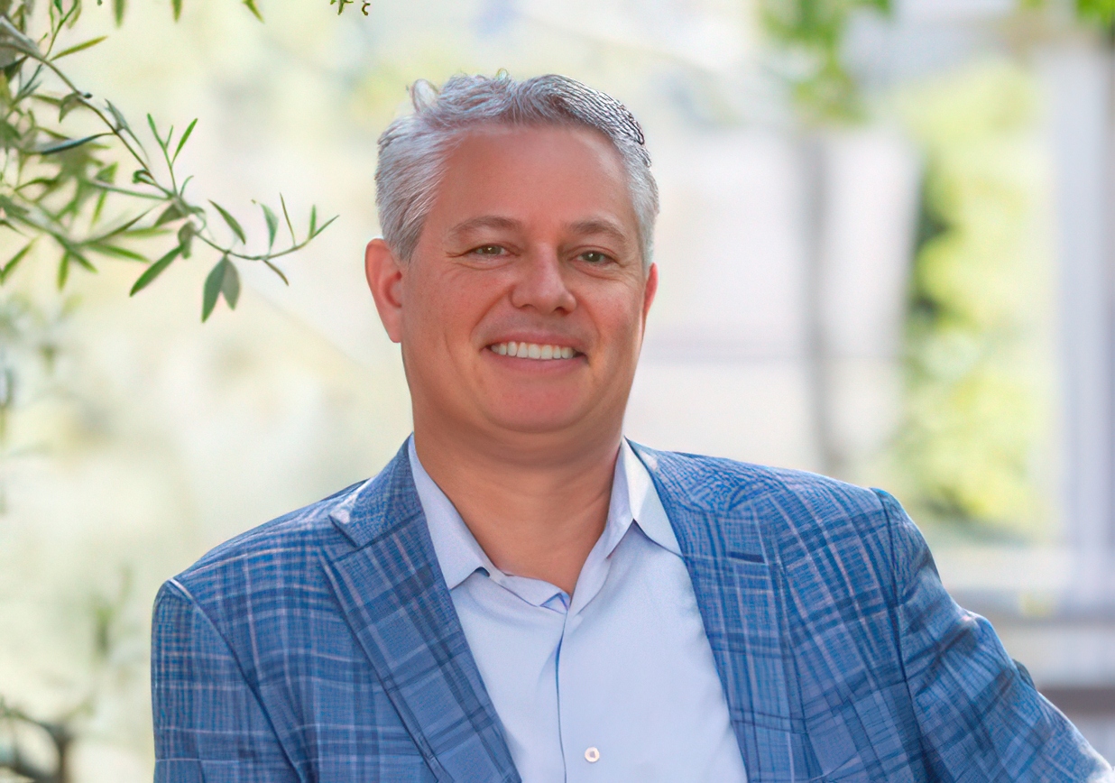 A smiling man with gray hair in a blue plaid suit jacket and light blue shirt, outdoors with blurred greenery in the